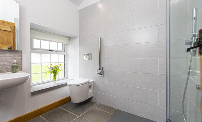 A modern bathroom featuring a walk-in rainfall shower, wall-mounted toilet, large frosted window with flowers, and light grey tiles with natural slate